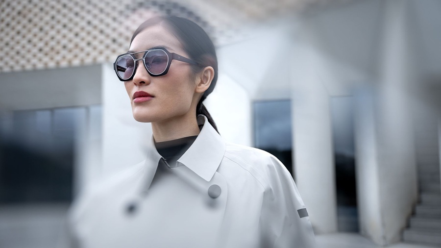Femme portant des lunettes de soleil Blackfin One Sun à montures en titane géométriques hexagonales et verres dégradés, vêtue d'un trench-coat blanc sur fond architectural flou