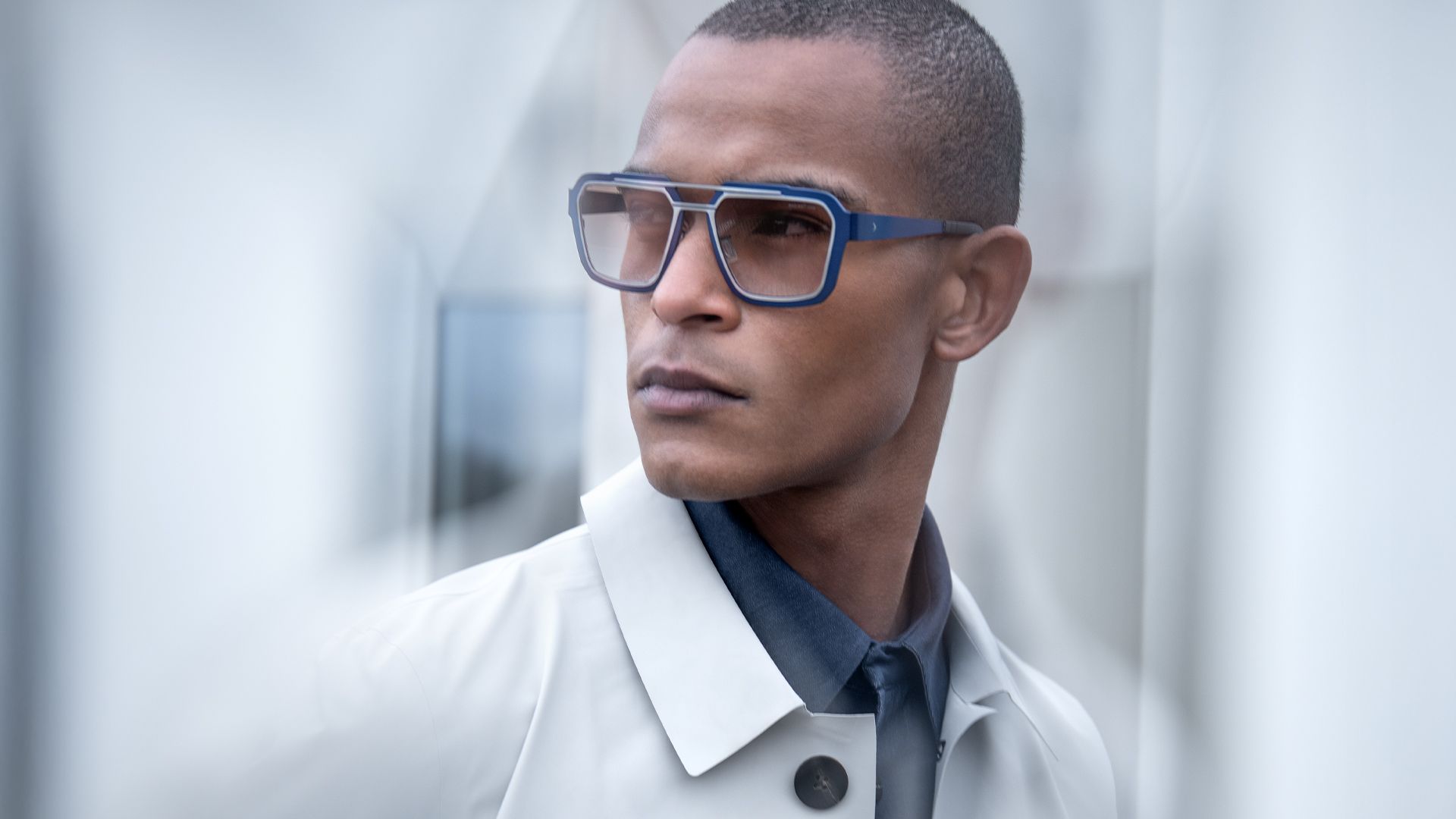 Man wearing Blackfin One Sun Iron Harbor sunglasses in bold blue square titanium frames with a shape-within-a-shape design, styled with a light grey trench coat