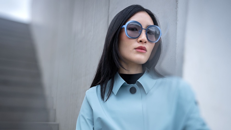 Woman wearing Blackfin One Sun Sandy Hook sunglasses in soft blue round titanium frames with gradient lenses, paired with a pale blue trench coat against a concrete wall