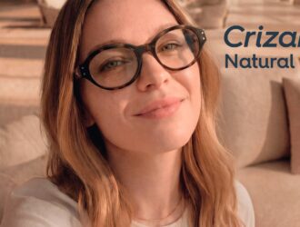 A woman wearing tortoiseshell glasses with Crizal® Natural Look™ anti-reflective lenses smiles warmly in a cozy indoor setting, with the Crizal® Natural Look™ logo displayed in the upper right corner.