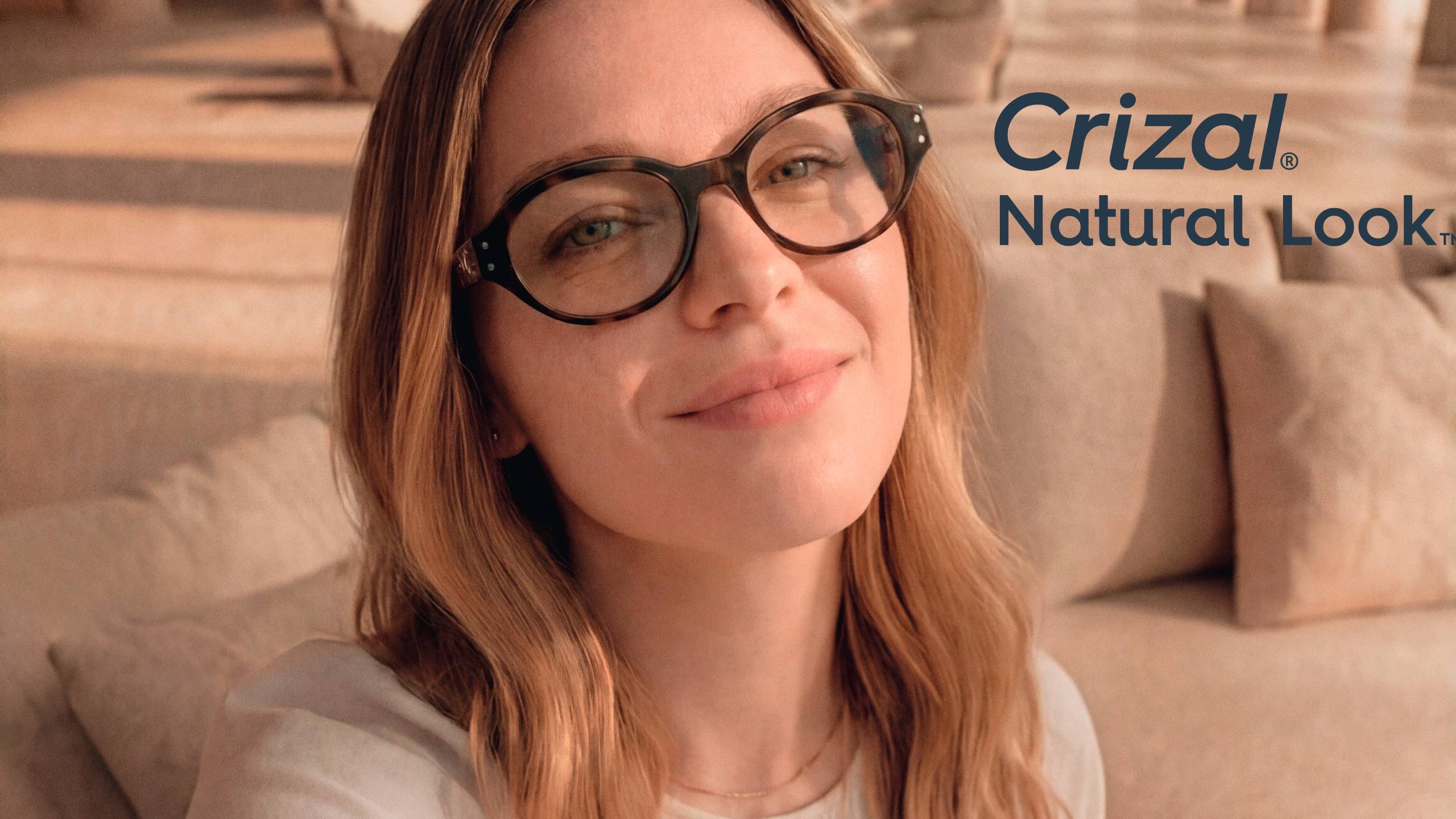 A woman wearing tortoiseshell glasses with Crizal® Natural Look™ anti-reflective lenses smiles warmly in a cozy indoor setting, with the Crizal® Natural Look™ logo displayed in the upper right corner.