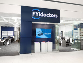 Exterior view of an FYidoctors retail clinic location inside a shopping mall, featuring a navy blue storefront with illuminated logo, a digital display screen showing a promotional offer, and eyeglass frames visible on display walls inside.