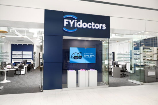 Exterior view of an FYidoctors retail clinic location inside a shopping mall, featuring a navy blue storefront with illuminated logo, a digital display screen showing a promotional offer, and eyeglass frames visible on display walls inside.