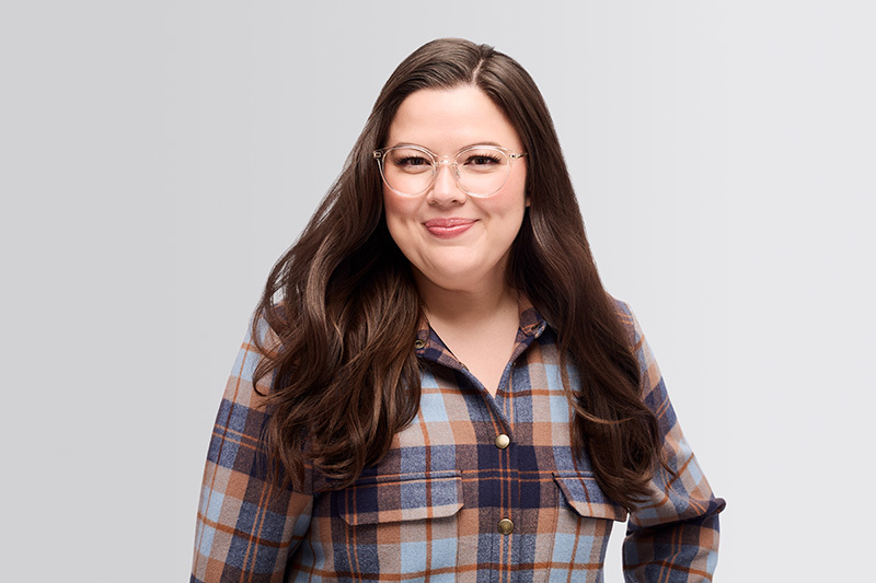 French: Hilary MacMillan souriante, portant des lunettes rondes rose clair et une chemise à carreaux bleu, brun et marine, sur un fond gris clair.