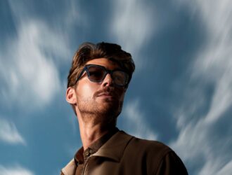 Un homme portant des lunettes à monture foncée avec des verres photochromiques teintés se tient en extérieur sous un ciel bleu dramatique strié de nuages blancs effilés. Les verres apparaissent activés à la lumière du soleil, illustrant les performances d'assombrissement en extérieur des verres HOYA Sensity® 3.