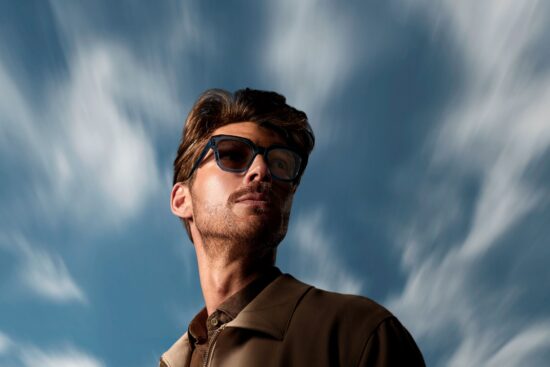 A man wearing dark-framed glasses with tinted photochromic lenses stands outdoors against a dramatic blue sky streaked with wispy white clouds. The lenses appear activated in the sunlight, demonstrating HOYA Sensity® 3's outdoor darkening performance.