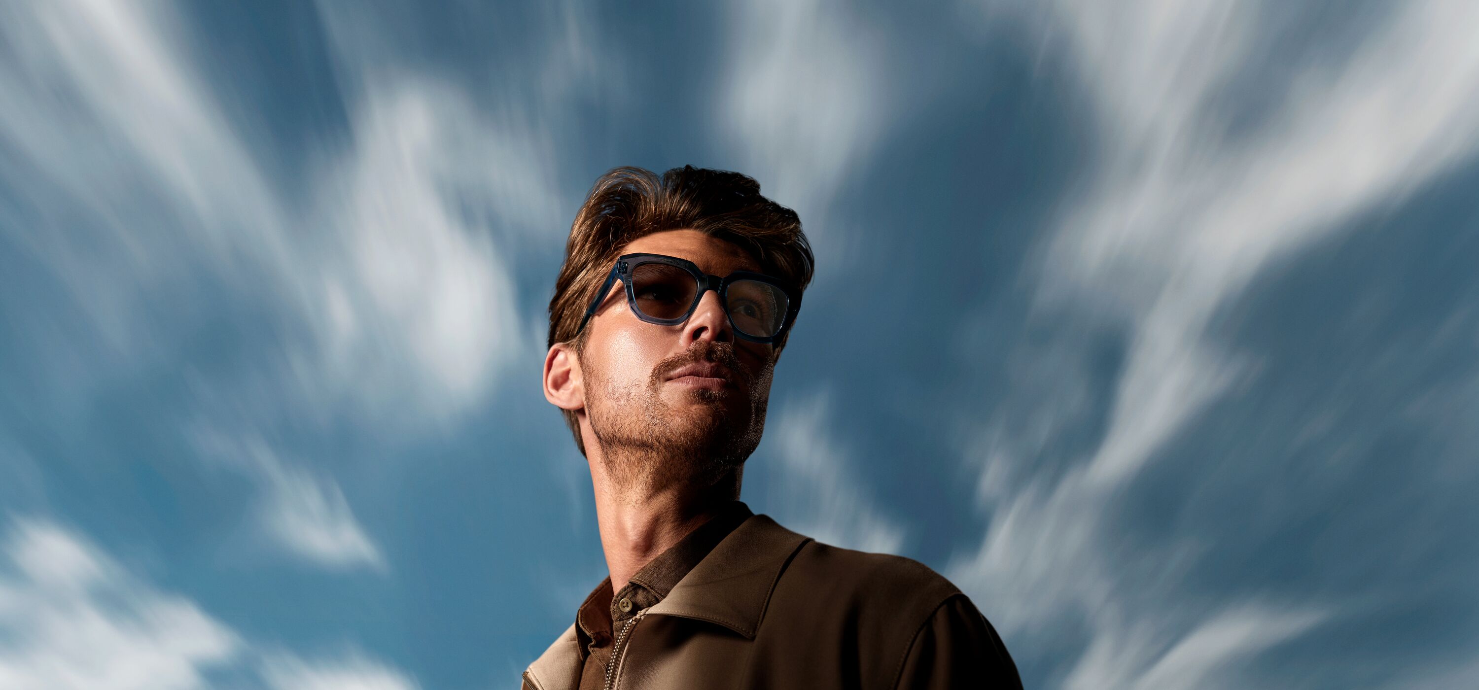 A man wearing dark-framed glasses with tinted photochromic lenses stands outdoors against a dramatic blue sky streaked with wispy white clouds. The lenses appear activated in the sunlight, demonstrating HOYA Sensity® 3's outdoor darkening performance.