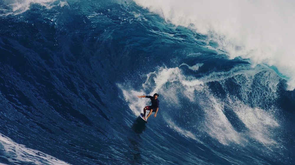 Kai Lenny surfant une vague massive, vue en plongée, avec de l'écume blanche déferlant derrière lui