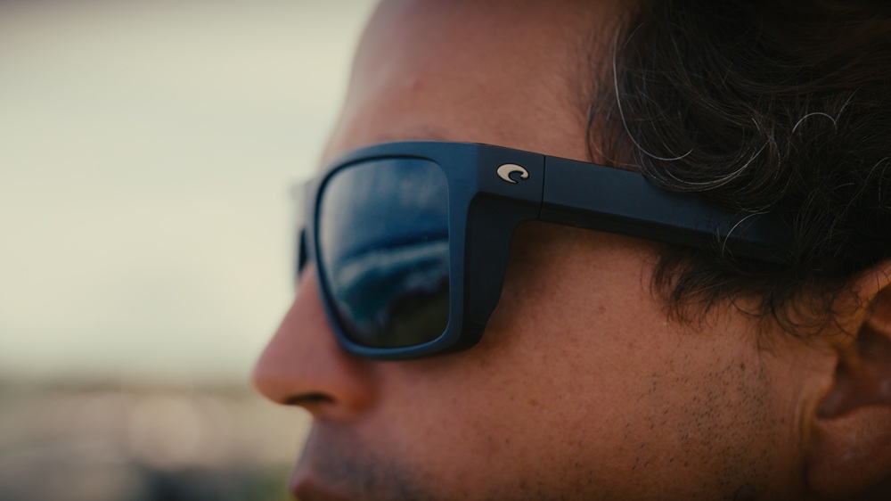 Profil rapproché de Kai Lenny portant les lunettes de soleil Costa Lido aux verres bleus miroir, avec le reflet de l'océan dans les verres