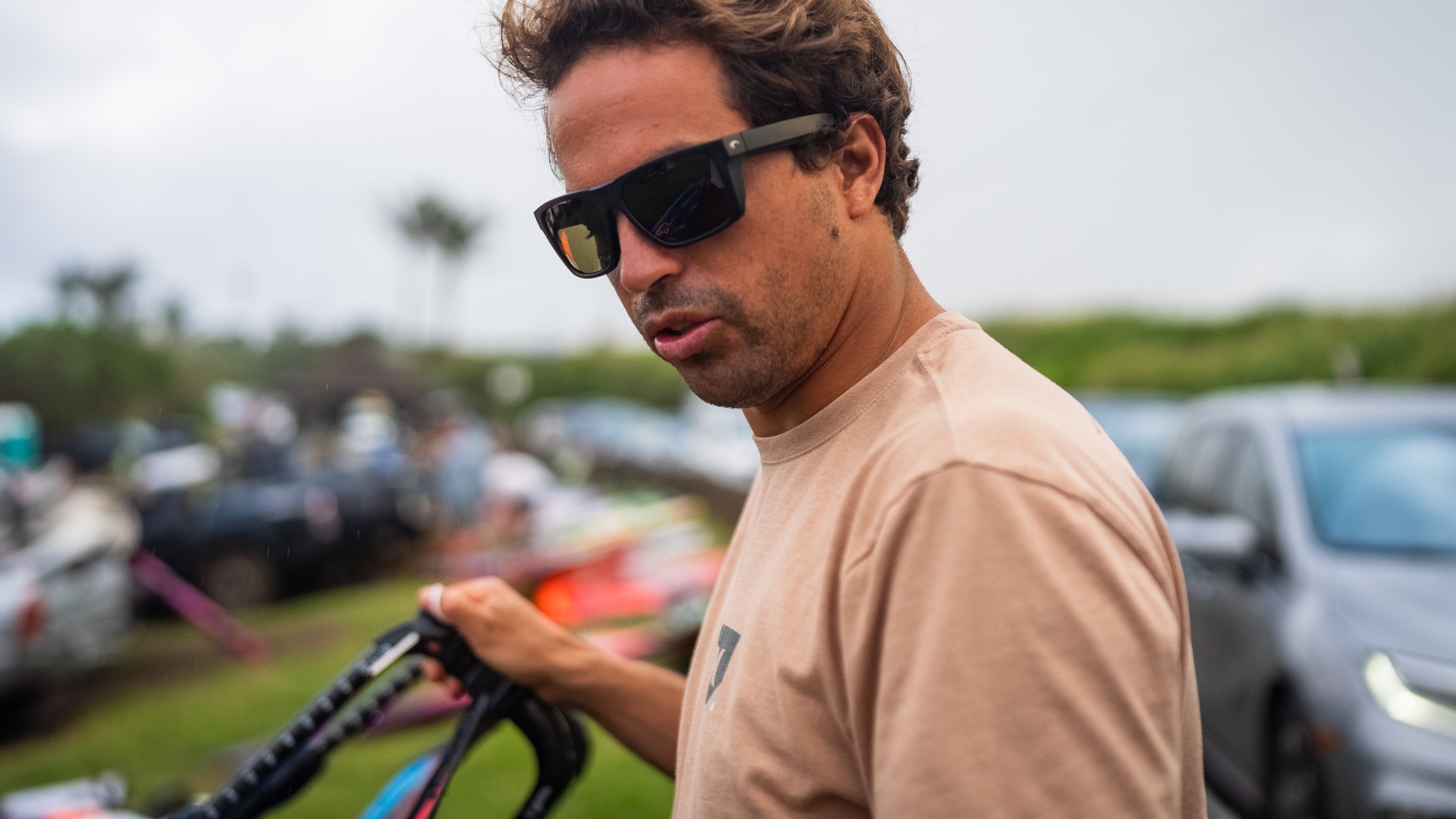 Kai Lenny portant des lunettes de soleil Costa aux verres ambrés, tenant du matériel de sports nautiques lors d'un événement en plein air avec une foule en arrière-plan