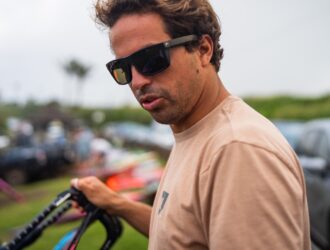 Kai Lenny wearing Costa sunglasses with amber lenses, holding water sports equipment at an outdoor event with a crowd in the background
