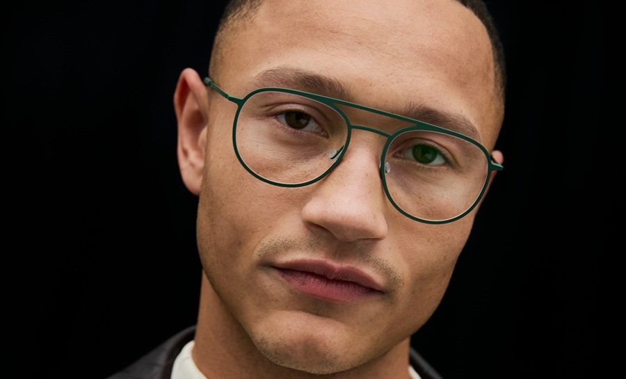 Un homme portant des lunettes aviateur à double pont en vert foncé, vêtu d'une chemise crème texturée et d'une veste en cuir noir embossé façon crocodile, photographié sur fond sombre.