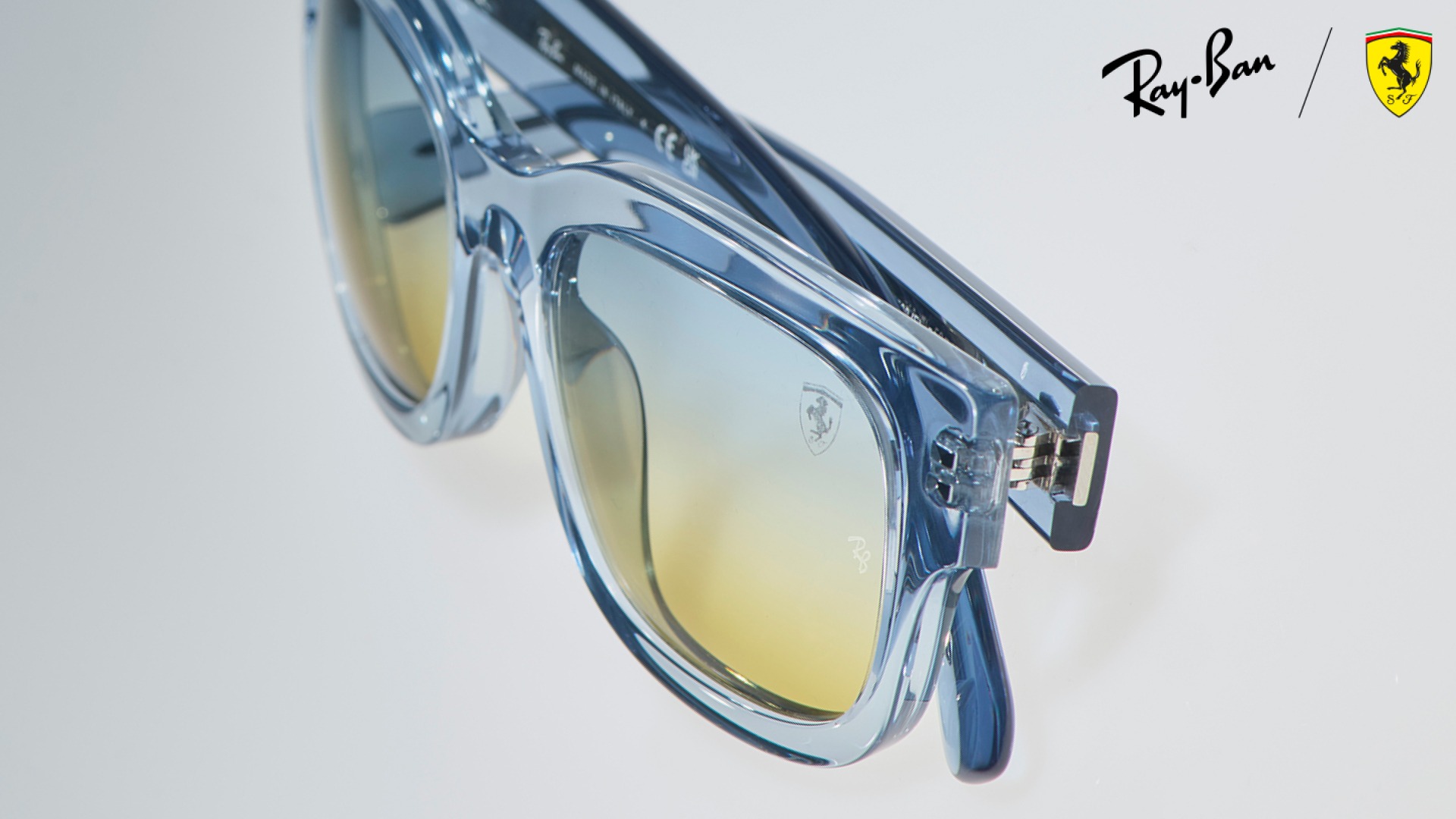 Ray-Ban x Scuderia Ferrari RB2228M sunglasses in transparent blue acetate with a blue-to-yellow gradient lens and Scuderia Ferrari shield logo, shown in close-up detail against a light background.