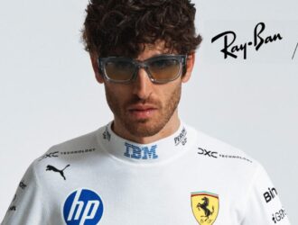 Male model wearing Ray-Ban x Scuderia Ferrari sunglasses in transparent blue frames, dressed in a white Scuderia Ferrari HP race suit, holding a red racing jacket, against a light grey background.