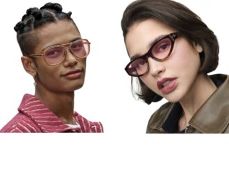 Two models wearing Transitions® Color Touch™ lenses in the Ruby color. Left model wears gold aviator frames with pink-rose tinted lenses and a striped pink jacket. Right model wears tortoiseshell cat-eye frames with rose-gradient lenses and an olive leather jacket.