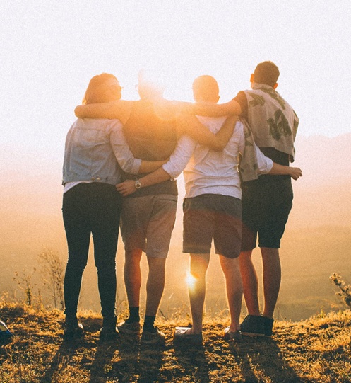 Four friends standing together with arms around each other, looking toward a bright sunset over a hazy landscape.