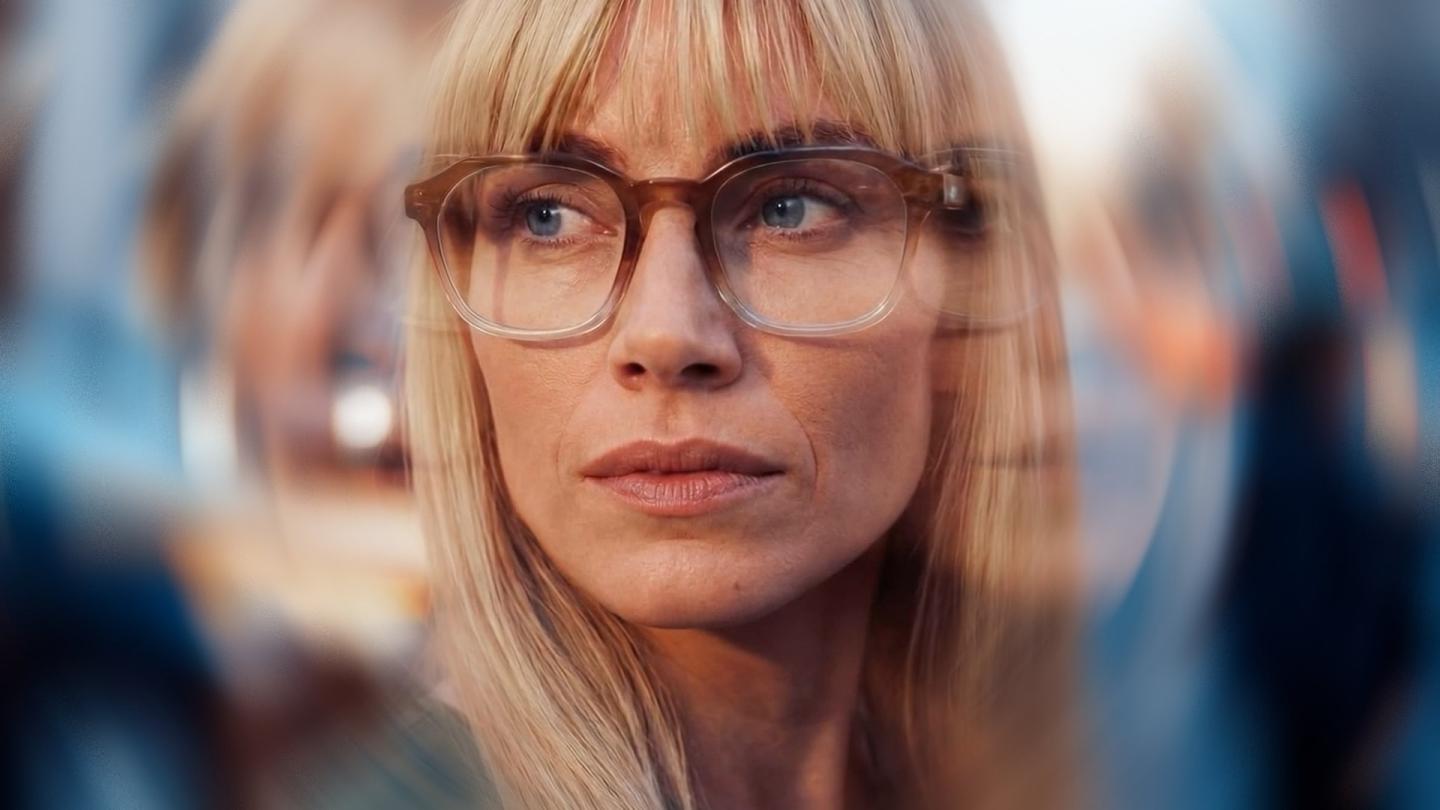 Une femme portant des lunettes à monture écaille regarde attentivement devant elle, entourée de reflets flous évoquant un environnement animé et visuellement stimulant