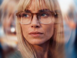 A woman wearing tortoiseshell frame glasses looks attentively ahead, surrounded by blurred reflections suggesting a busy, visually stimulating environment