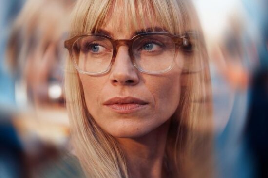 A woman wearing tortoiseshell frame glasses looks attentively ahead, surrounded by blurred reflections suggesting a busy, visually stimulating environment