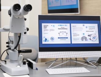 A slit lamp biomicroscope and a desktop computer displaying an ophthalmology clinic consultation webpage, set on an examination desk in an eye care office.