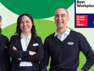 Trois employés de Specsavers en uniforme marine souriant et riant, aux côtés du badge Great Place To Work® Meilleurs employeurs™ Canada 2026, sur un fond vert et vert clair.