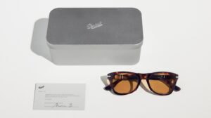Lunettes de soleil Persol en écailles présentées à côté d'un étui en métal brossé minimaliste et d'une carte d'authenticité signée.