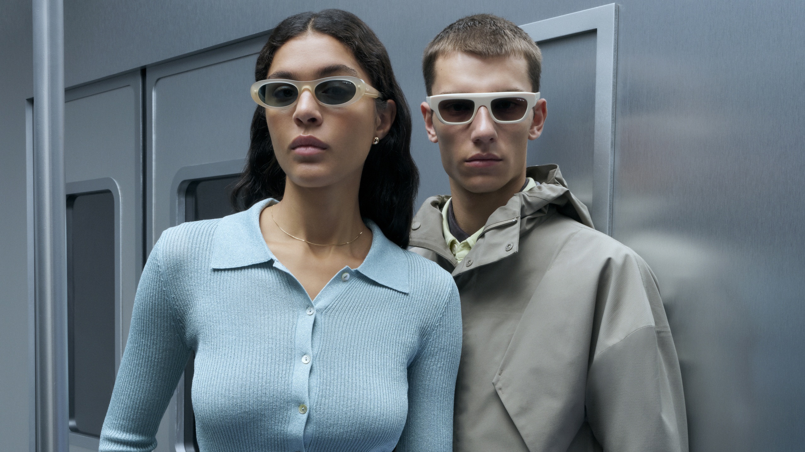 Two models wearing the SS26 collection: a woman in narrow oval-shaped sunglasses and a man in chunky, rectangular white frames.