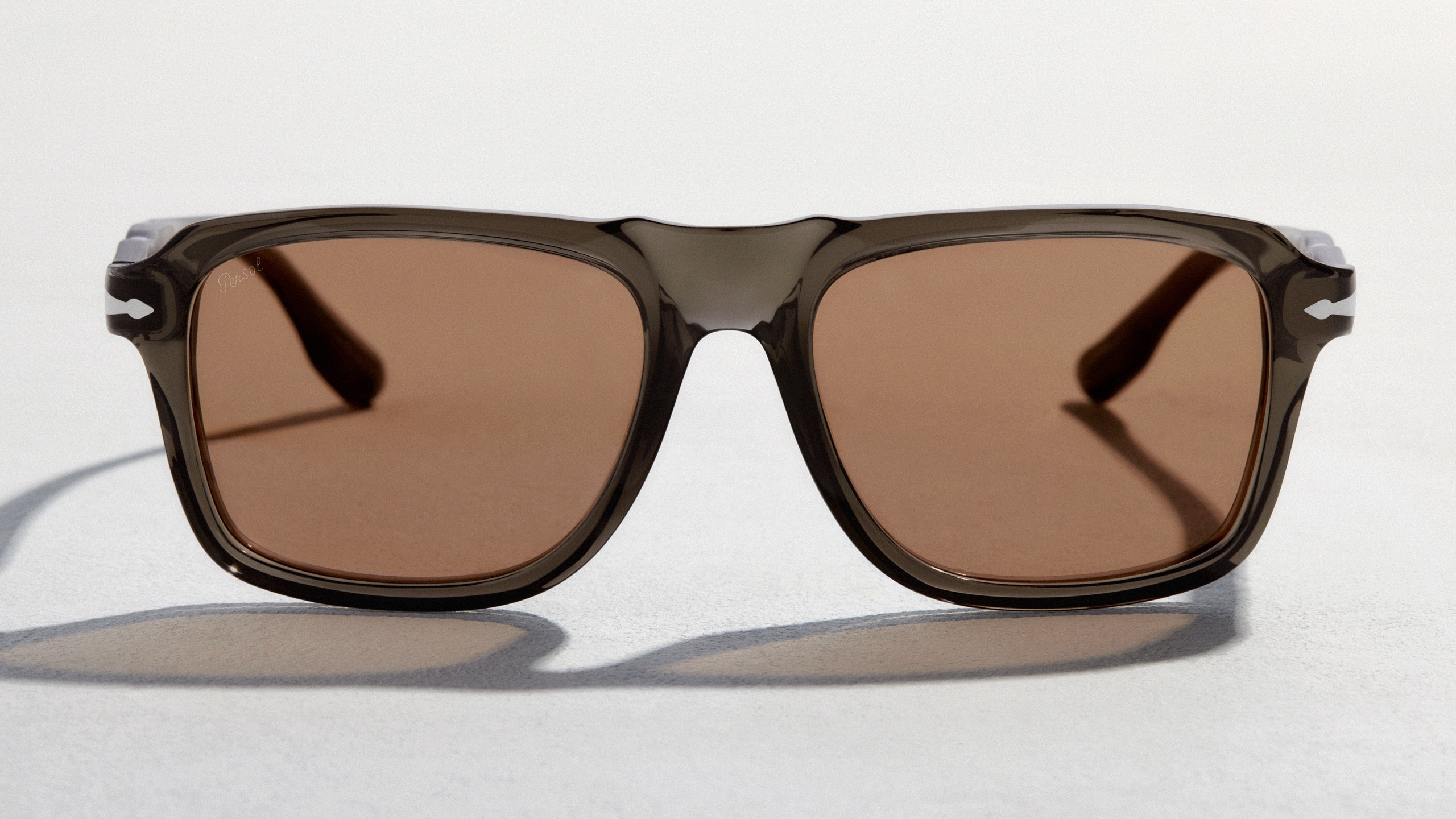 Front-facing view of Persol sunglasses featuring a smoke-grey transparent frame and warm brown lenses