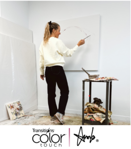 Artist AnnLo stands in her bright white studio, painting the first strokes of a large blank canvas on the wall. She is dressed in a white sweater and black pants, holding a paintbrush mid-gesture. A paint-covered work table stands beside her. At the bottom of the image, the co-branded logos of Transitions® Color Touch™ and AnnLo are displayed side by side.