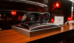 A close-up shot of the "Wayfarer Puffer Diamond" eyewear displayed on a sleek, metallic silver pedestal atop a wooden surface. The black frames are entirely encrusted with shimmering crystals or small diamonds, with the Ray-Ban logo visible in gold on the temple. The background is dark with soft red bokeh highlights.
