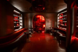 The interior of Ray-Ban House showing a continuous, cinematic design with satin metal curved walls and deep red floors. The room features long, built-in leather banquette seating and small, round metallic pedestal tables. Shelves recessed into the walls display eyewear alongside various objects under soft lighting.