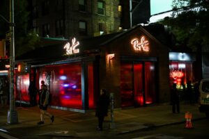 An exterior wide shot of the two-story townhouse at the corner of Prince and Lafayette. The brick building features a glowing "Ray-Ban HOUSE" neon sign on its gabled roofline. Large windows are saturated with red light and (RED) branding, while people walk by on the New York City sidewalk at night.