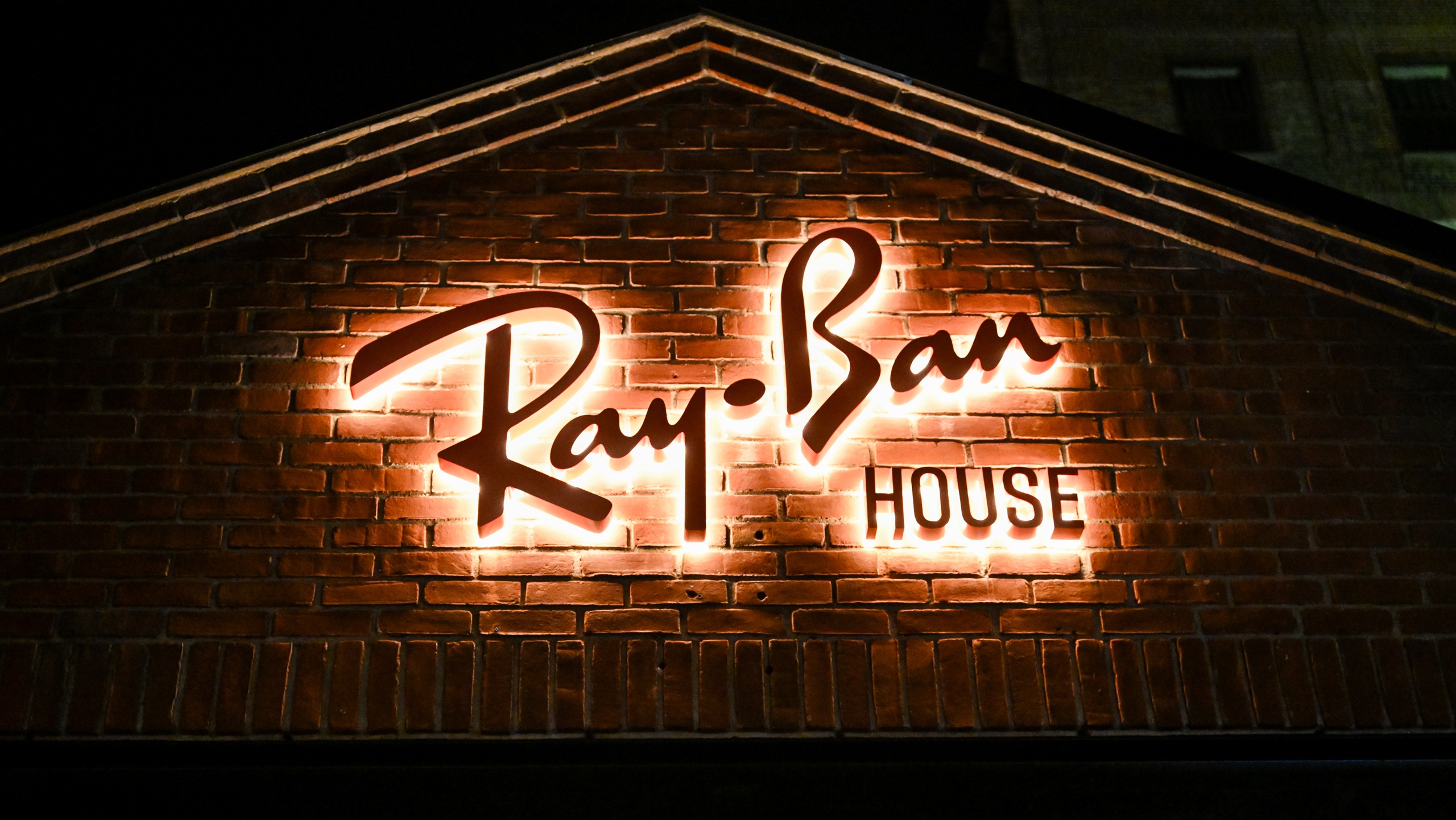 Un plan rapproché de l'enseigne « Ray-Ban HOUSE » fixée sur un mur de briques rouges classiques. L'écriture cursive « Ray-Ban » et le mot « HOUSE » sont rétroéclairés, créant un halo blanc chaleureux contre la brique sombre durant la nuit.