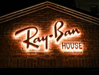 A tight architectural shot of the "Ray-Ban HOUSE" signage mounted on a classic red brick wall. The "Ray-Ban" script and the word "HOUSE" are backlit, creating a warm, white halo effect against the dark brick at night.