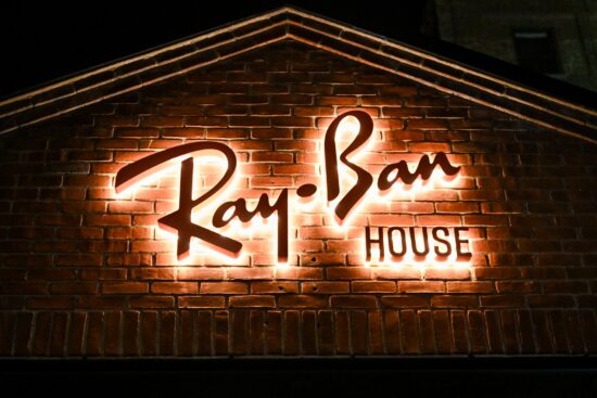A tight architectural shot of the "Ray-Ban HOUSE" signage mounted on a classic red brick wall. The "Ray-Ban" script and the word "HOUSE" are backlit, creating a warm, white halo effect against the dark brick at night.
