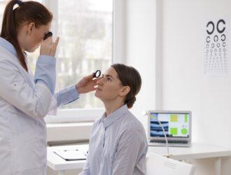 Une ophtalmologiste en blouse blanche utilise une lentille portative pour examiner l'œil d'une patiente assise dans un bureau médical lumineux. À l'arrière-plan, un ordinateur portable affiche des examens oculaires à côté d'une échelle de Snellen fixée au mur.
