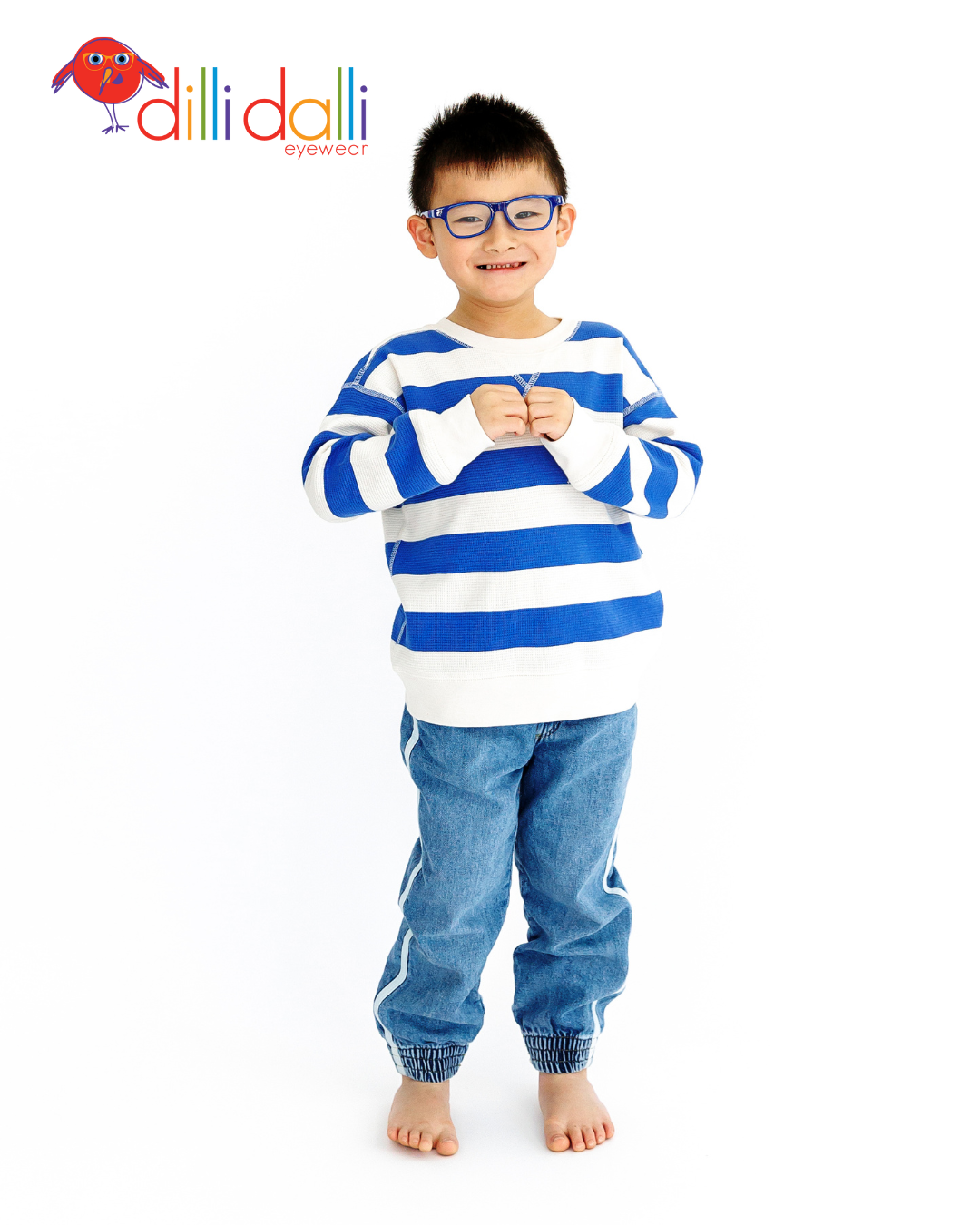 A young boy smiling and wearing blue Dilli Dalli Hero glasses, a blue and white striped sweater, and denim joggers.