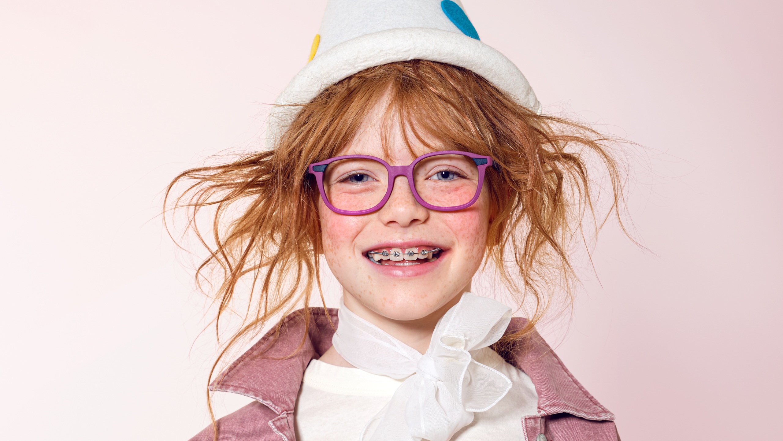 Une jeune fille rousse souriante avec un appareil dentaire, portant des lunettes LOOKKINO TRI-X violettes et un chapeau blanc ludique.
