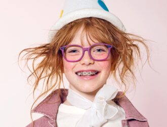 A smiling young girl with red hair and braces wearing vibrant purple LOOKKINO TRI-X glasses and a playful white hat.
