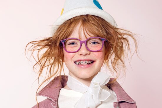 A smiling young girl with red hair and braces wearing vibrant purple LOOKKINO TRI-X glasses and a playful white hat.