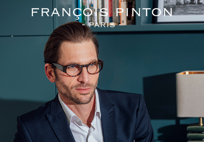 A man wearing tortoiseshell François Pinton glasses and a navy blue suit jacket, sitting in front of a bookshelf.