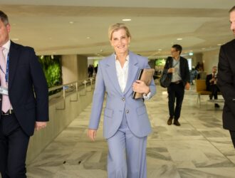 HRH The Duchess of Edinburgh, Global Ambassador for the IAPB, smiling and walking through a bright, modern hallway at the World Health Assembly. She is wearing a light blue suit and carrying a tan clutch and laptop, accompanied by officials in suits.