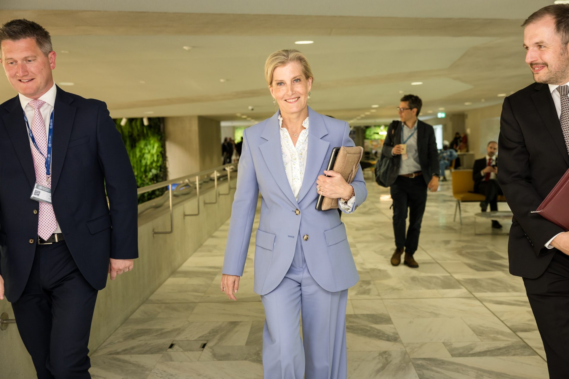 S.A.R. la duchesse d'Édimbourg, ambassadrice mondiale de l'IAPB, marchant en souriant dans un couloir moderne lors de l'Assemblée mondiale de la Santé. Elle porte un tailleur bleu clair et tient une pochette ainsi qu'un ordinateur portable, entourée de collaborateurs en costume.