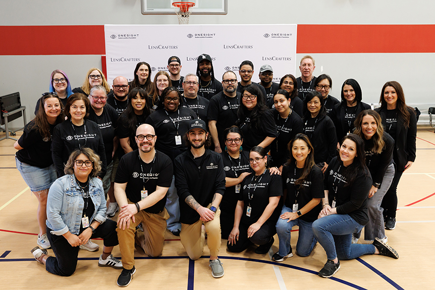 Un groupe d'environ 30 bénévoles de LensCrafters et de la Fondation OneSight EssilorLuxottica posent pour une photo d'équipe sur un terrain de basket. Ils portent des t-shirts noirs assortis aux logos des organisations et se tiennent devant une bannière blanche.