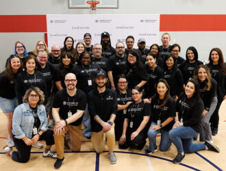 A large group of approximately 30 LensCrafters and OneSight EssilorLuxottica Foundation volunteers pose for a team photo on a basketball court. They are wearing matching black branded t-shirts and standing in front of a white backdrop featuring the organization logos.