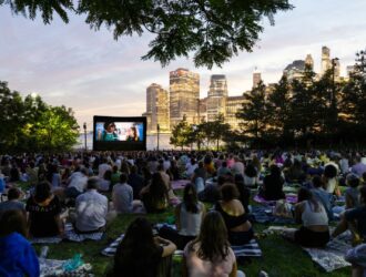 Une grande foule de New-Yorkais installés sur des couvertures et des tapis de pique-nique sur la pelouse de Brooklyn Bridge Park regardent une projection de film en plein air au crépuscule. Un grand écran de cinéma gonflable lumineux occupe le premier plan, tandis que la skyline illuminée de Manhattan et un ciel aux teintes rosées forment un arrière-plan spectaculaire. Des arbres encadrent la scène par le haut. Cet événement s'inscrit dans le cadre de la série cinématographique estivale Movies With A View, présentée en partenariat avec Persol.