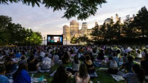 A large crowd of New Yorkers sit on blankets and picnic mats on the grass at Brooklyn Bridge Park, watching an outdoor film screening at dusk. A large inflatable movie screen glows in the foreground, while the illuminated Manhattan skyline and a pink-hued twilight sky form a stunning backdrop. Trees frame the scene from above. Part of the Movies With A View summer film series, presented in partnership with Persol.
