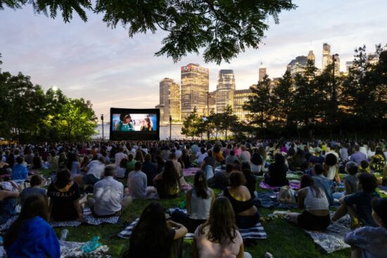 A large crowd of New Yorkers sit on blankets and picnic mats on the grass at Brooklyn Bridge Park, watching an outdoor film screening at dusk. A large inflatable movie screen glows in the foreground, while the illuminated Manhattan skyline and a pink-hued twilight sky form a stunning backdrop. Trees frame the scene from above. Part of the Movies With A View summer film series, presented in partnership with Persol.