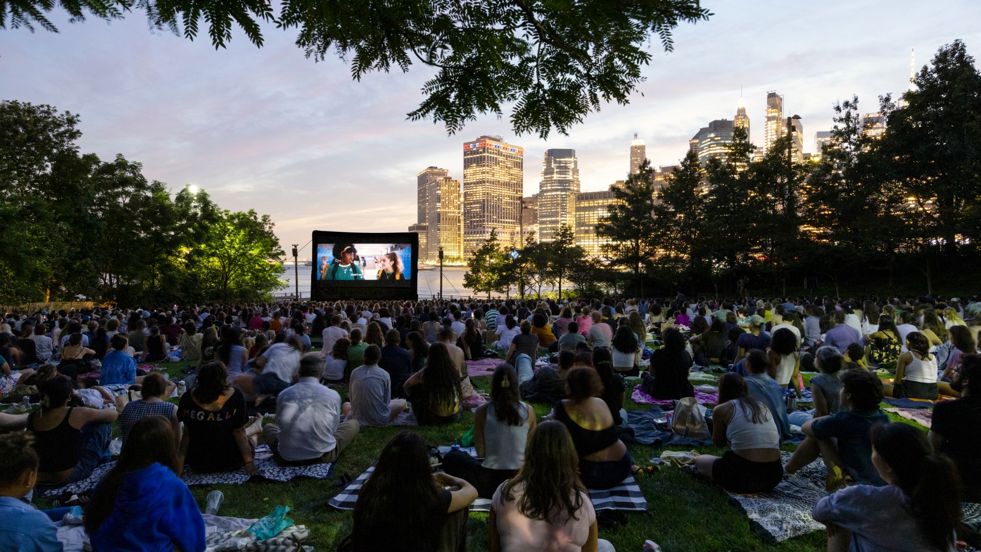 A large crowd of New Yorkers sit on blankets and picnic mats on the grass at Brooklyn Bridge Park, watching an outdoor film screening at dusk. A large inflatable movie screen glows in the foreground, while the illuminated Manhattan skyline and a pink-hued twilight sky form a stunning backdrop. Trees frame the scene from above. Part of the Movies With A View summer film series, presented in partnership with Persol.