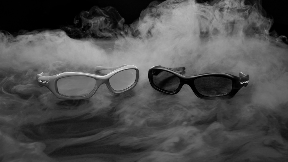 Deux paires de lunettes Oakley MUZM FatCat, une claire et une foncée, présentées ensemble dans un décor dramatique rempli de fumée.
